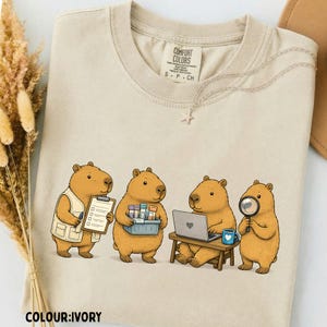 May include: An ivory t-shirt featuring a graphic of four brown capybaras engaged in scientific tasks. One holds a clipboard, another carries test tubes, one works on a laptop, and the fourth uses a magnifying glass. The shirt has a Comfort Colors label.