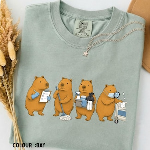 May include: A sage green t-shirt featuring a graphic of four brown capybaras performing various cleaning and medical tasks. One capybara holds a clipboard, another mops, one carries a cleaning bucket, and the fourth wears a medical mask.