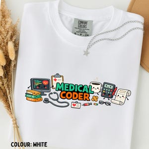 Op de afbeelding: Een wit t-shirt met een kleurrijke afbeelding en de tekst MEDICAL CODER. Het ontwerp bevat cartoonillustraties van een computermonitor, een stethoscoop, een rekenmachine, een koffiekopje en boeken met de labels ICD 10 en CPT.