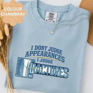 Może przedstawiać: Niebieska koszulka typu t-shirt w kolorze chambray z grafiką przedstawiającą zdjęcie rentgenowskie kości oraz napis I DONT JUDGE APPEARANCES I JUDGE FRACTURES. Koszulka ma okrągły dekolt i białą metkę wewnętrzną z widoczną nazwą marki Comfort Colors.