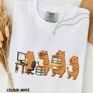 May include: A white t-shirt featuring a graphic of four brown capybaras engaged in office and study tasks. One sits at a computer desk, another wears glasses while holding a document, a third carries a stack of books, and the fourth holds a paper.