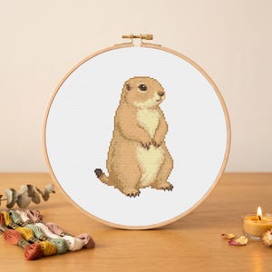 May include: A circular wooden embroidery hoop containing a cross-stitch pattern of a standing prairie dog. The prairie dog is rendered in shades of tan and cream, featuring a small black eye and a dark-tipped tail, centered on white fabric.