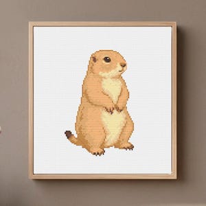 May include: A square framed cross-stitch artwork featuring a pixelated prairie dog in shades of tan and cream. The animal is depicted in a standing position against a white grid background, enclosed within a light-colored wooden frame.