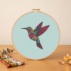 May include: A circular wooden embroidery hoop containing a cross-stitch pattern of a hummingbird on a light blue fabric background. The hummingbird features a floral design with shades of pink, red, green, and yellow within its wings and body.