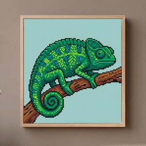 May include: A square framed cross-stitch art piece featuring a pixel art style chameleon in shades of green, yellow, and blue. The chameleon is perched on a brown branch against a light blue background, all enclosed within a light wood frame.