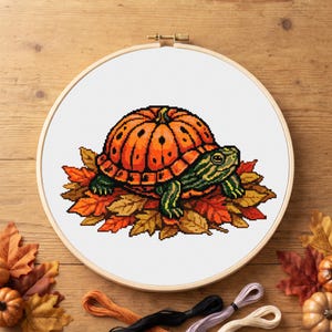 May include: A cross-stitch embroidery project in a circular wooden hoop featuring a pixel-art style turtle with a bright orange pumpkin shell, resting on a bed of colorful autumn leaves, all stitched onto a white fabric background.