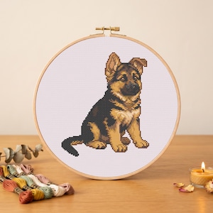 May include: A cross-stitch embroidery project featuring a German Shepherd puppy in black and tan thread, displayed in a round wooden hoop. The design is centred on white Aida fabric, showcasing the puppy in a seated position with alert ears.