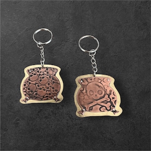 May include: Two wooden keychains with metal skull designs. One keychain features a pattern of skulls, while the other displays a skull and crossbones. Both have silver key rings and chains, with decorative metal accents.