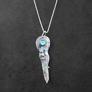 May include: A silver-toned pendant necklace with a key-shaped design. The pendant features intricate detailing and a small, round, light blue stone. The necklace hangs on a delicate silver chain, set against a dark background.