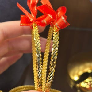 May include: Two thin, circular gold bangles featuring a twisted, rope-like texture. Each bangle is adorned with a small, bright red fabric ribbon tied at the top. The metallic surface of the bangles has a polished, reflective finish.