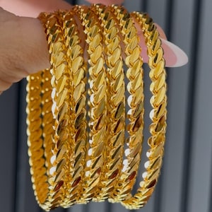 Puede incluir: Un conjunto de cinco brazaletes dorados, cada uno con un patrón repetitivo de eslabones trenzados y texturizados. Los brazaletes tienen una superficie pulida y reflectante que capta la luz, creando un aspecto metálico brillante en los aros.
