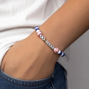 Op de afbeelding: Een kralenarmband met blauwe en witte ronde kralen, twee kralen met een honkbalpatroon, een goudkleurige metalen kraal en witte letterkralen die 99 JUDGE spellen. De armband is gemaakt met een flexibel elastisch koord.