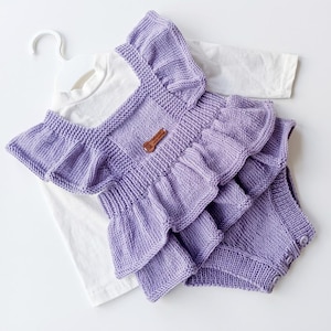 Puede incluir: Un pelele de punto color lavanda con tirantes con volantes y un diseño de falda superpuesta, sobre una camiseta blanca de manga larga. El pelele incluye una pequeña etiqueta rectangular marrón en el pecho y tres botones en la abertura de la pierna.