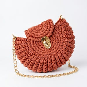 Pode incluir: Uma bolsa de crochê cor de ferrugem em formato semicircular com um padrão de malha texturizada, uma aba dobrável e um fecho de pressão em metal dourado polido. A bolsa inclui uma alça de ombro de corrente de metal dourado a condizer.