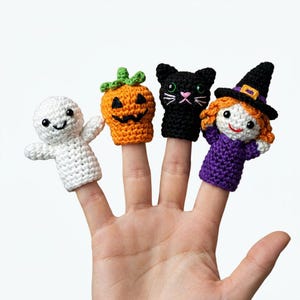 May include: A set of four crocheted finger puppets featuring a white ghost, an orange pumpkin with a green stem, a black cat with green eyes, and a character wearing a purple dress and a black witch hat with a yellow band on the brim.