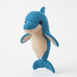 May include: A handcrafted crochet dolphin toy featuring a vibrant blue body with a contrasting beige underbelly. The plush figure has a rounded head, two side fins, a dorsal fin, and a tail fin, all worked in a textured yarn with black eyes.