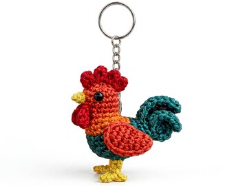 May include: A crochet rooster keychain featuring a red comb and wattle, an orange head and wing, a dark green body, and a teal tail. The rooster has a yellow beak and feet, black bead eyes, and is attached to a silver metal split ring keychain.