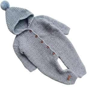 May include: A light blue knitted one-piece jumpsuit featuring a hood topped with a matching pom-pom. The garment has long sleeves, ribbed cuffs, and a front button closure with five small brown buttons. A small rectangular tag is on the left leg.