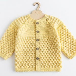 May include: A pale yellow long-sleeve knit cardigan featuring a textured bobble stitch pattern on the body and sleeves, a smooth ribbed yoke, a round neckline, and five dark brown buttons down the center front, displayed on a wooden hanger.