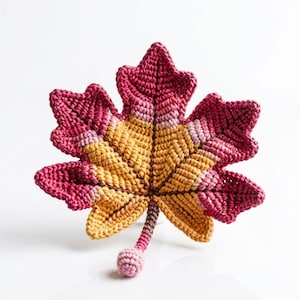 May include: A crochet maple leaf ornament featuring a gradient color pattern that transitions from deep magenta at the outer edges to a warm golden yellow in the center. The leaf has a textured surface and a small stem with a round ball at the end.