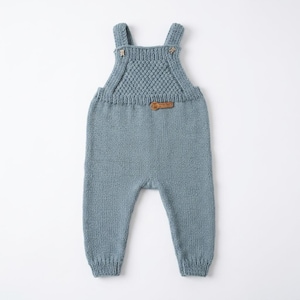May include: A dusty blue knitted overall featuring a textured honeycomb pattern on the chest, two shoulder straps with small bear-shaped buttons, and a rectangular brown leather-like brand tag centred below the chest panel.