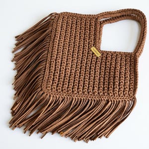 May include: A brown crocheted handbag featuring a square body with a textured ribbed pattern, a single integrated handle, and long fringe detailing along the side and bottom edges. A small rectangular tag labelled Handmade is attached.
