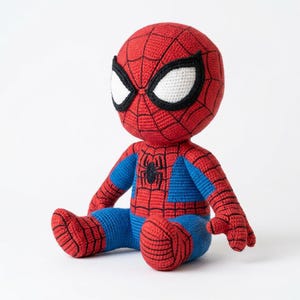Puede incluir: Una muñeca de Spider-Man tejida a ganchillo en posición sentada, con un cuerpo de hilo rojo y azul con detalles de telaraña negros. La figura tiene grandes ojos blancos delineados en negro y un pequeño emblema de araña negro en el pecho.