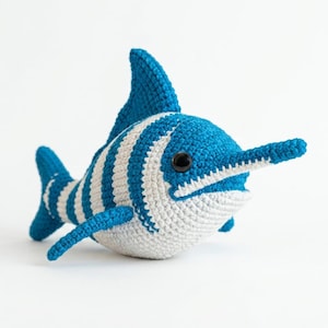 May include: A crocheted stuffed toy shaped like a swordfish with a blue and white striped pattern. The toy features a long, pointed snout, a prominent dorsal fin, a small tail fin, and two side fins. It has a single black plastic eye on its side.