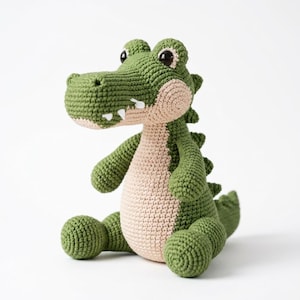 Puede incluir: Un juguete de cocodrilo tejido a ganchillo hecho a mano con un cuerpo verde texturizado, vientre y hocico de color beige claro. El peluche tiene grandes ojos de plástico negro, pequeños dientes de fieltro blanco y espinas verdes en el lomo.