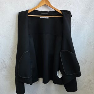 May include: A black open-front jacket with long sleeves and two large rounded patch pockets on the front. The garment features a white label at the inner neckline with the text Acne Studios printed in black. The jacket is displayed on a wooden hanger.