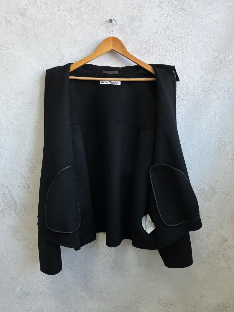 May include: A black open-front jacket with long sleeves and two large rounded patch pockets on the front. The garment features a white label at the inner neckline with the text Acne Studios printed in black. The jacket is displayed on a wooden hanger.