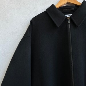 May include: A black zip-up jacket with a pointed collar and long sleeves. The garment features a full-length front zipper closure and a visible brand label on the inner neck area that reads Acne Studios. The fabric has a textured, felted appearance.