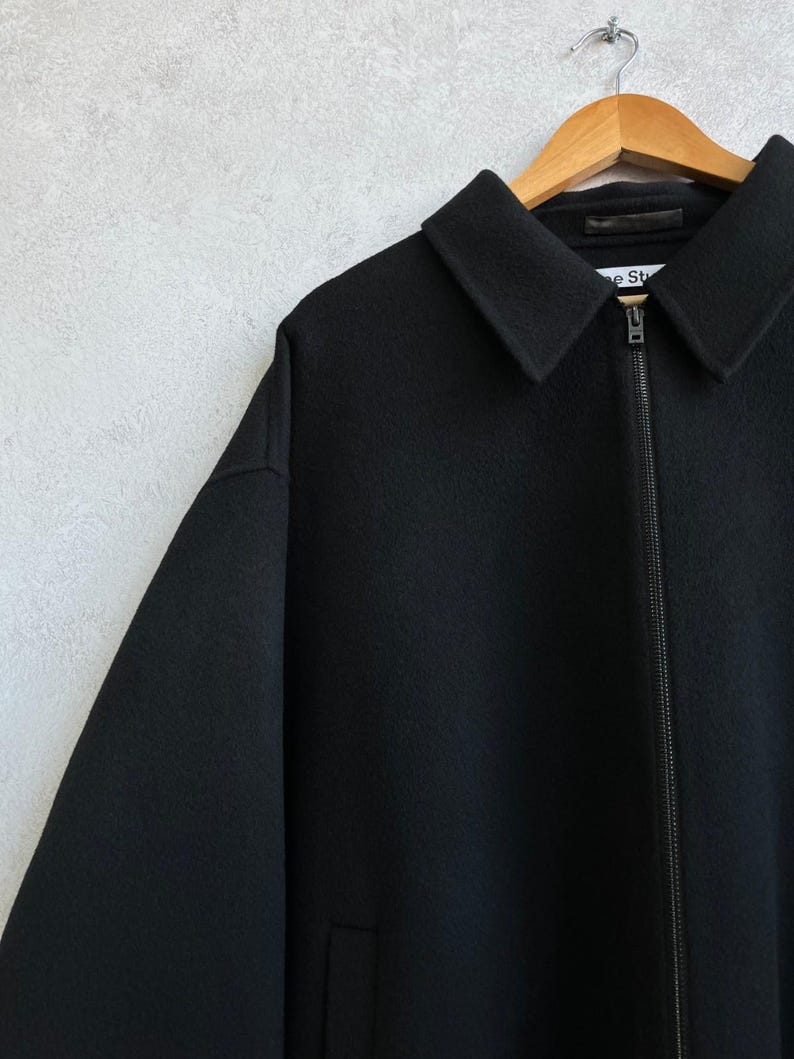 May include: A black zip-up jacket with a pointed collar and long sleeves. The garment features a full-length front zipper closure and a visible brand label on the inner neck area that reads Acne Studios. The fabric has a textured, felted appearance.