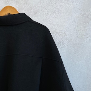 May include: A solid black jacket featuring a classic collar, a horizontal seam across the upper back, and long sleeves. The garment is displayed on a wooden hanger, showcasing a textured fabric surface and a structured, clean-cut design.