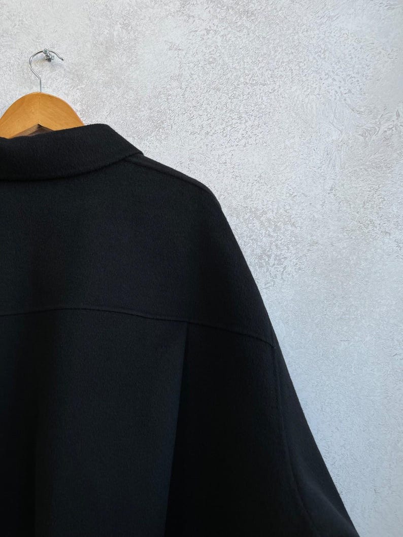 May include: A solid black jacket featuring a classic collar, a horizontal seam across the upper back, and long sleeves. The garment is displayed on a wooden hanger, showcasing a textured fabric surface and a structured, clean-cut design.