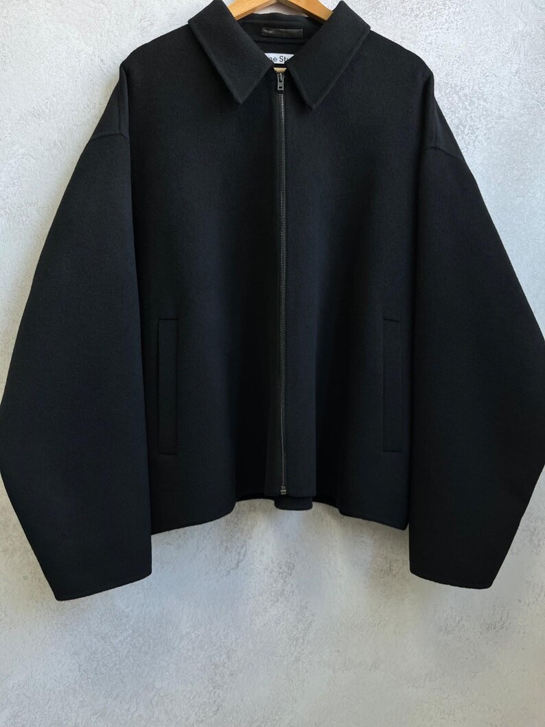 May include: A solid black jacket featuring a pointed collar, a full-length front zipper closure, and two vertical slit pockets. The garment has long, wide sleeves and a straight hemline, with the brand label Acne Studios visible at the neck.