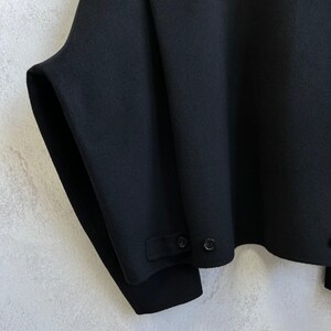 May include: A black jacket featuring a wide, structured silhouette with a horizontal seam across the upper chest. The hem includes a small tab detail secured with two black buttons, and the sleeves are wide with a clean, minimalist finish.