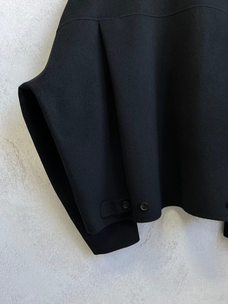 May include: A black jacket featuring a wide, structured silhouette with a horizontal seam across the upper chest. The hem includes a small tab detail secured with two black buttons, and the sleeves are wide with a clean, minimalist finish.
