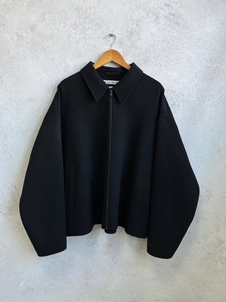 May include: A black zip-up jacket with a pointed collar, long sleeves, and two side pockets. The garment features a central zipper closure and a white interior label with the text Acne Studios, hanging on a wooden hanger against a textured wall.