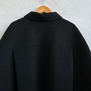 May include: A solid black jacket featuring a classic collar, a horizontal seam across the upper back, and a smooth, matte fabric texture. The garment is displayed on a wooden hanger, showcasing a clean, structured design with a simple, minimalist aesthetic.