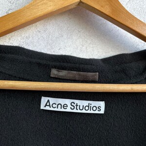 May include: A black garment with a soft, textured surface hanging on a wooden hanger. A white rectangular fabric label is sewn onto the interior neckline, featuring the black text Acne Studios in a clean, sans-serif font, with a smaller dark tag above.