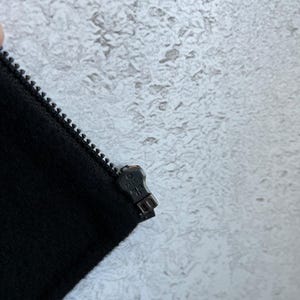 May include: A black fabric garment featuring a metal zipper track and a dark metal zipper pull embossed with the word TALON. The zipper pull is positioned at the top of the closed track, showing the metallic texture and the brand name clearly.