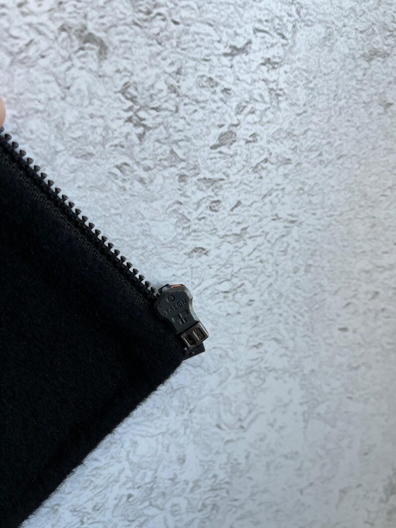 May include: A black fabric garment featuring a metal zipper track and a dark metal zipper pull embossed with the word TALON. The zipper pull is positioned at the top of the closed track, showing the metallic texture and the brand name clearly.