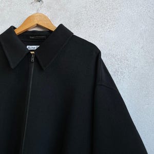 May include: A black zip-up jacket with a pointed collar and a front zipper closure. The interior label at the neckline displays the text Acne S. The garment is constructed from a solid black fabric and features long sleeves with a structured shoulder line.