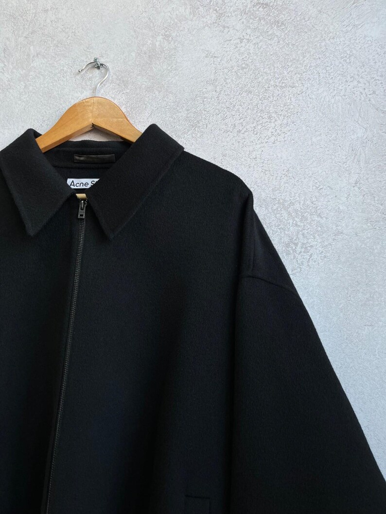 May include: A black zip-up jacket with a pointed collar and a front zipper closure. The interior label at the neckline displays the text Acne S. The garment is constructed from a solid black fabric and features long sleeves with a structured shoulder line.