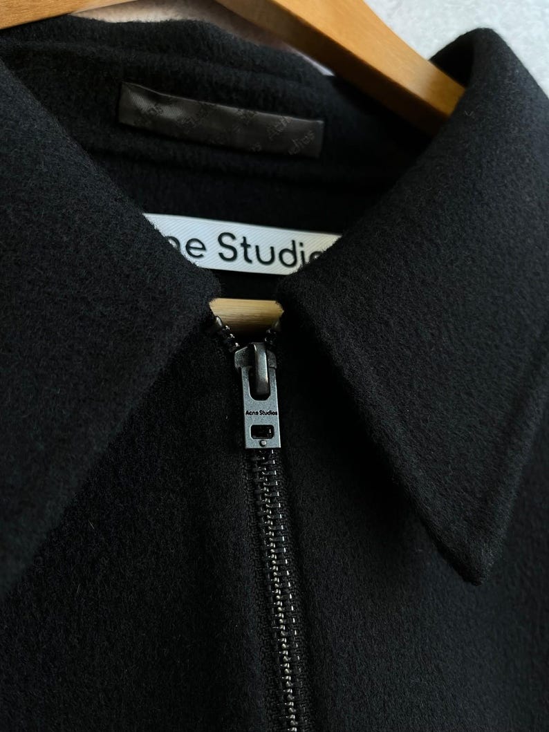May include: A black jacket with a pointed collar and a full-length front zipper. The zipper pull is metallic and features the text Acne Studios. A white interior label is visible at the neckline with the text Acne Studios printed in black.