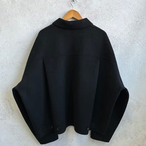 May include: A black jacket with a wide, boxy silhouette, a classic collar, and dropped shoulders. The garment features a horizontal back yoke, long sleeves, and a straight hemline with small button details at the lower sides, all displayed on a wooden hanger.