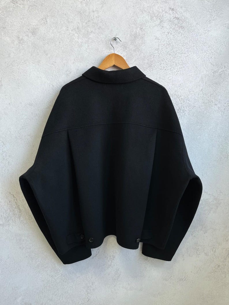 May include: A black jacket with a wide, boxy silhouette, a classic collar, and dropped shoulders. The garment features a horizontal back yoke, long sleeves, and a straight hemline with small button details at the lower sides, all displayed on a wooden hanger.