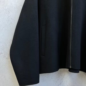 May include: A solid black jacket featuring a full-length front zipper closure, long sleeves, and a vertical welt pocket on the side. The garment is constructed from a dense, felted fabric with a smooth, matte texture and clean, straight seams.