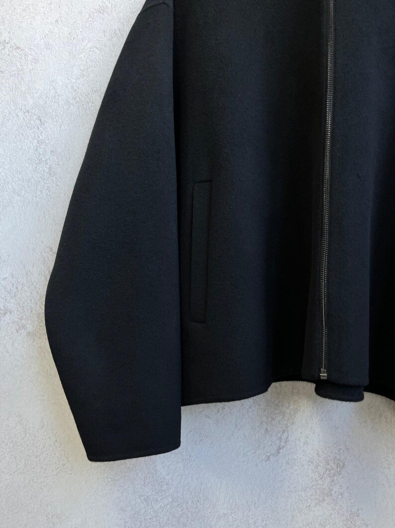 May include: A solid black jacket featuring a full-length front zipper closure, long sleeves, and a vertical welt pocket on the side. The garment is constructed from a dense, felted fabric with a smooth, matte texture and clean, straight seams.